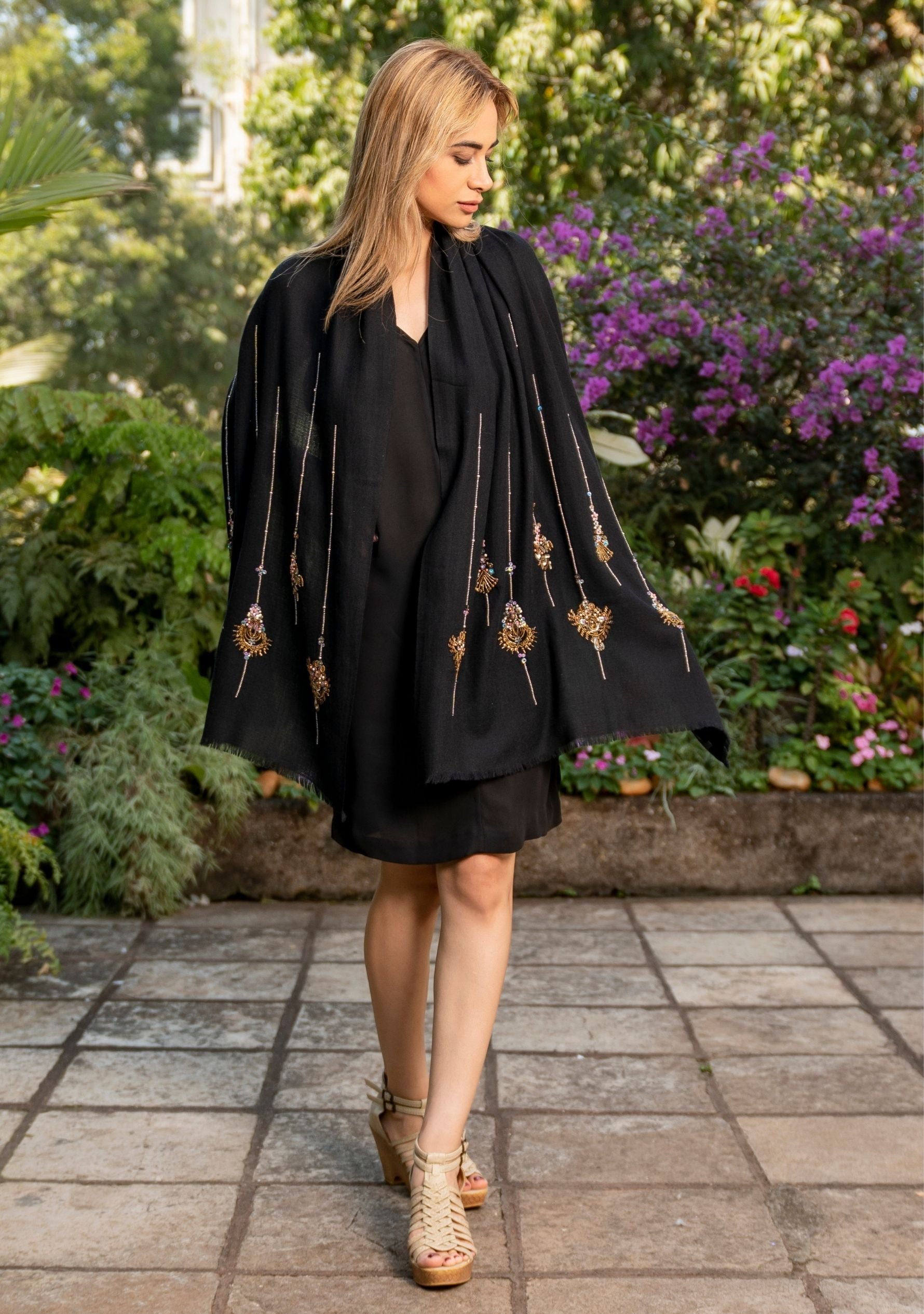 A woman in a black outfit with decorative elements walking outdoors on a paved path with greenery and flowers in the background. with Maneesha Ruia Black Scarf