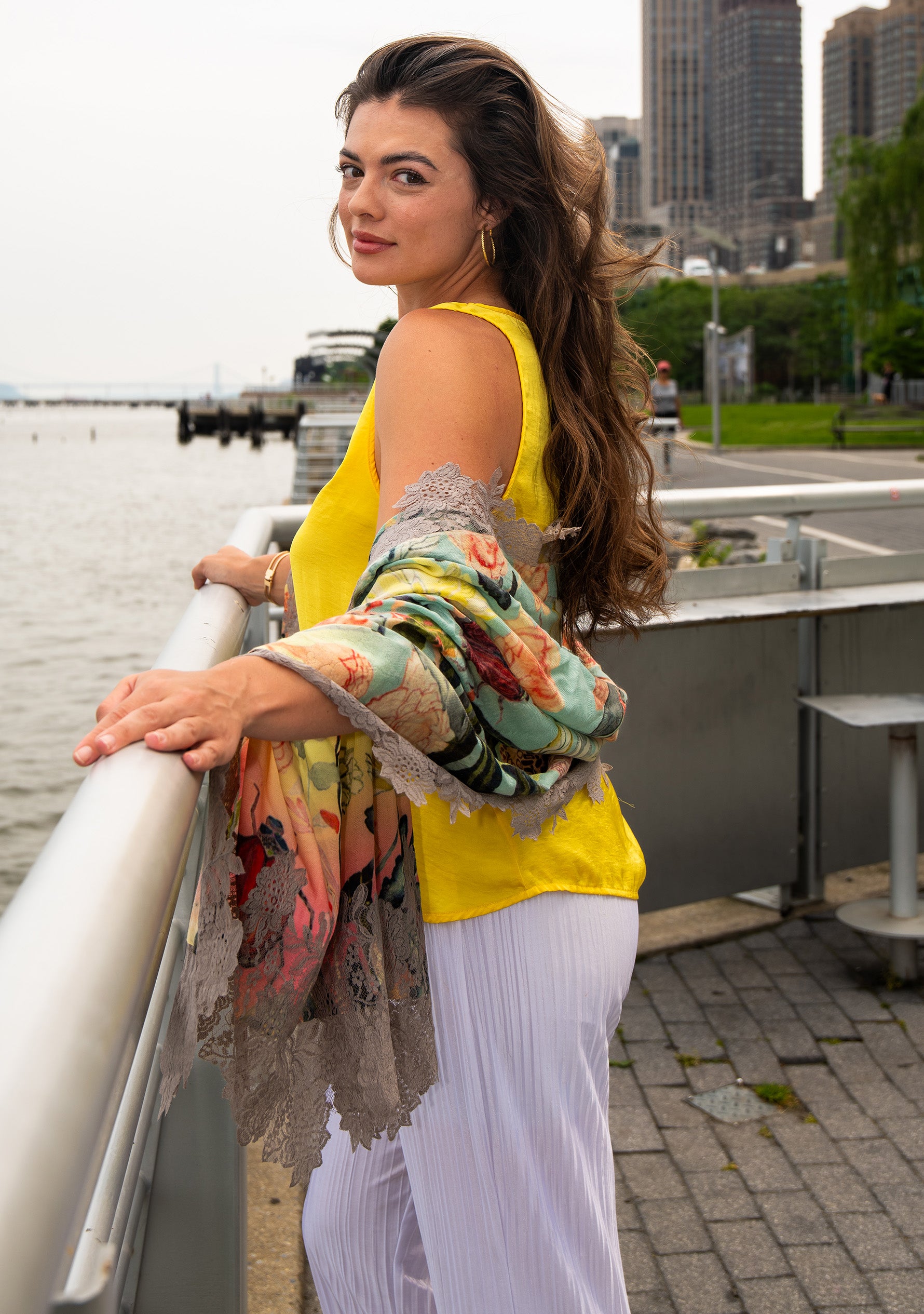 Woman in yellow top and white skirt with colorful MANEESHA RUIA SCARF leaning on a railing by a waterfront.