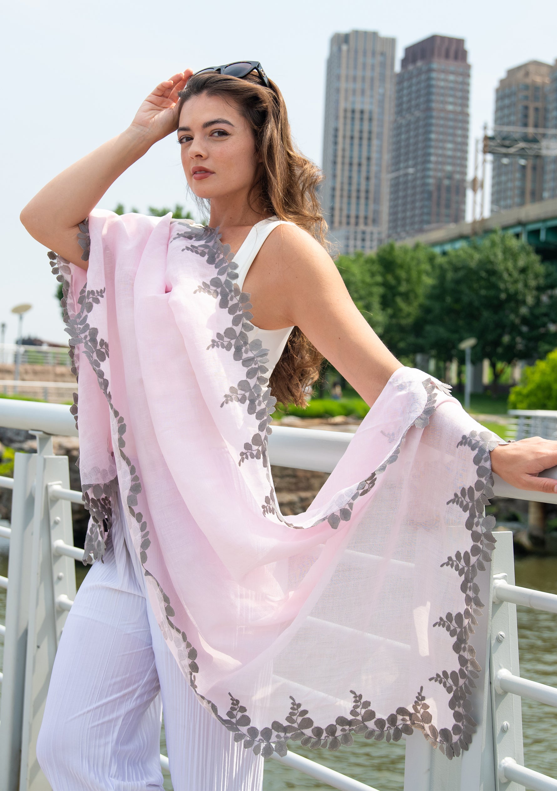 Woman holding a light pink scarf with gray floral patterns by a waterfront with buildings in the background
