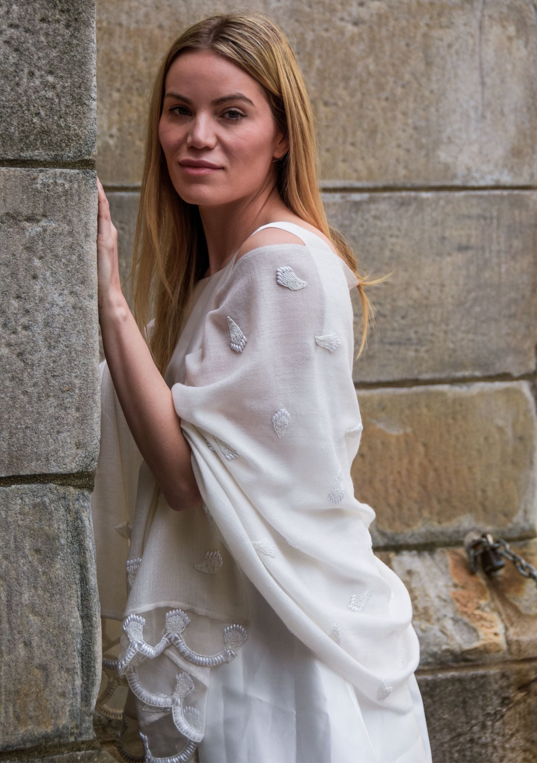 Woman wearing a white dress with lace details against a stone wall.