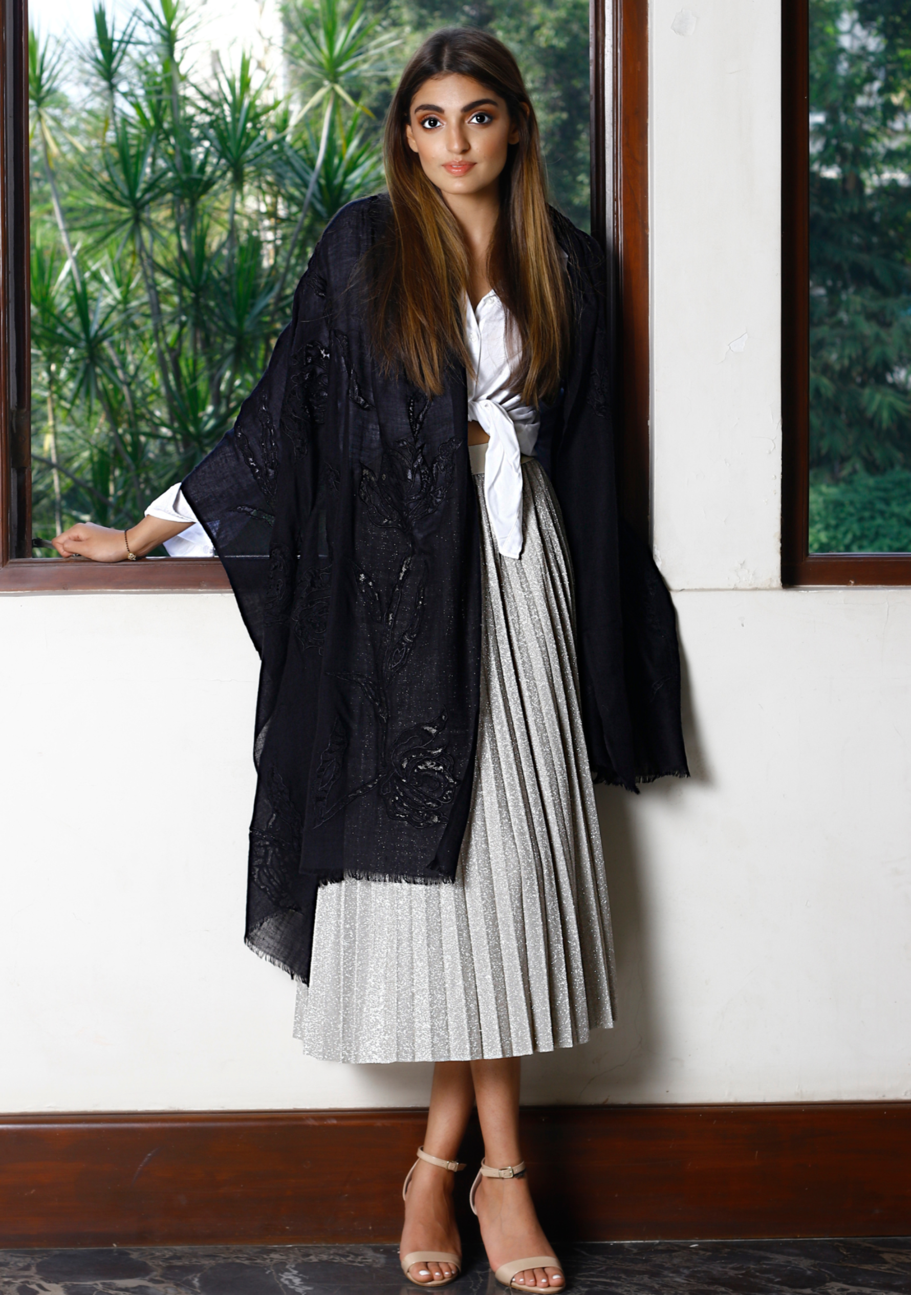 A woman wearing a black cashmere scarf with black rose lace applique, paired with a white and silver pleated dress, white blouse, and beige heels.