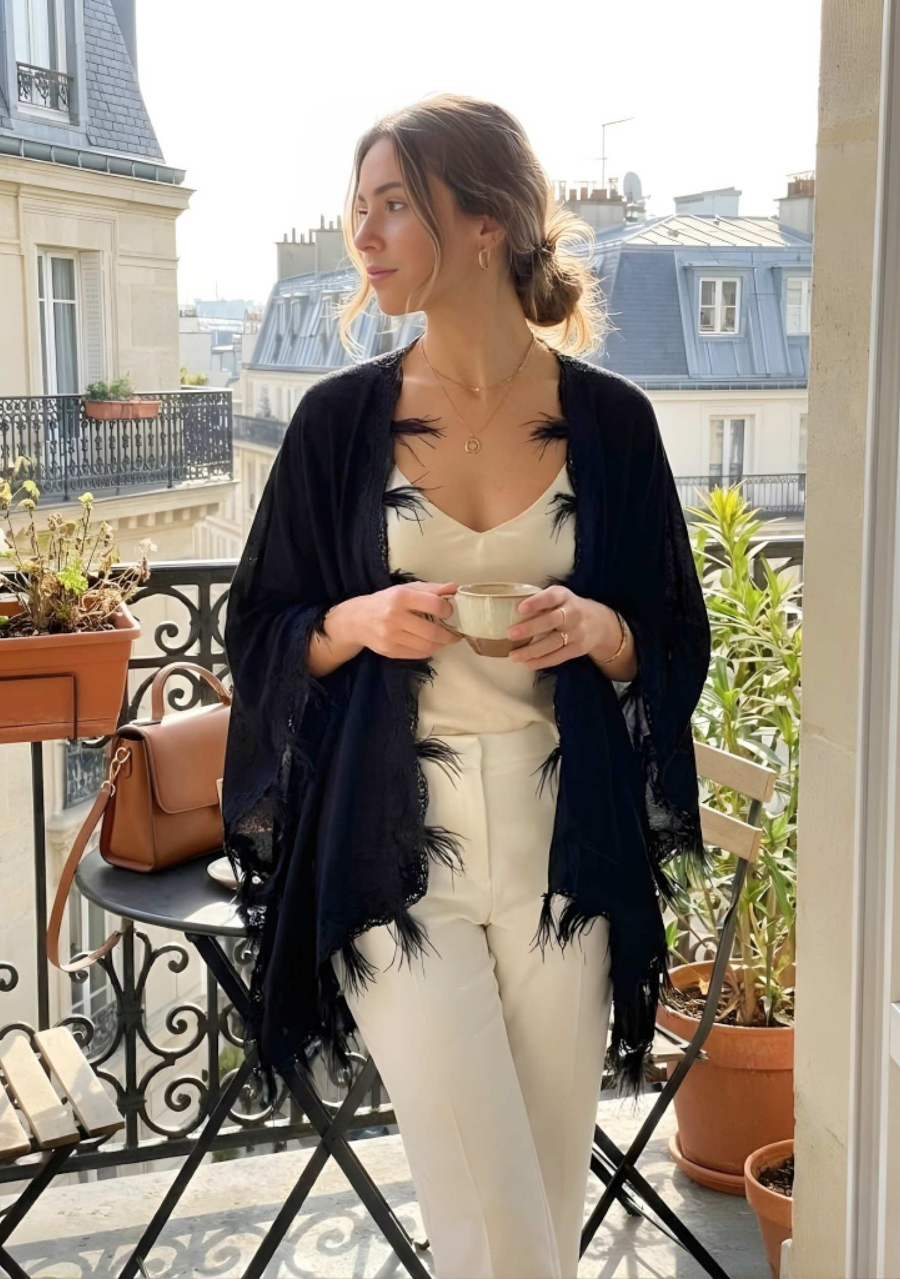 Woman standing on a balcony holding a cup, wearing a black cardigan and white pants.