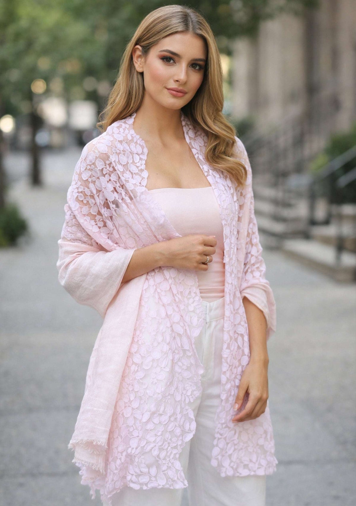 Woman wearing a light pink lace shawl over a white top and pants on a city street.