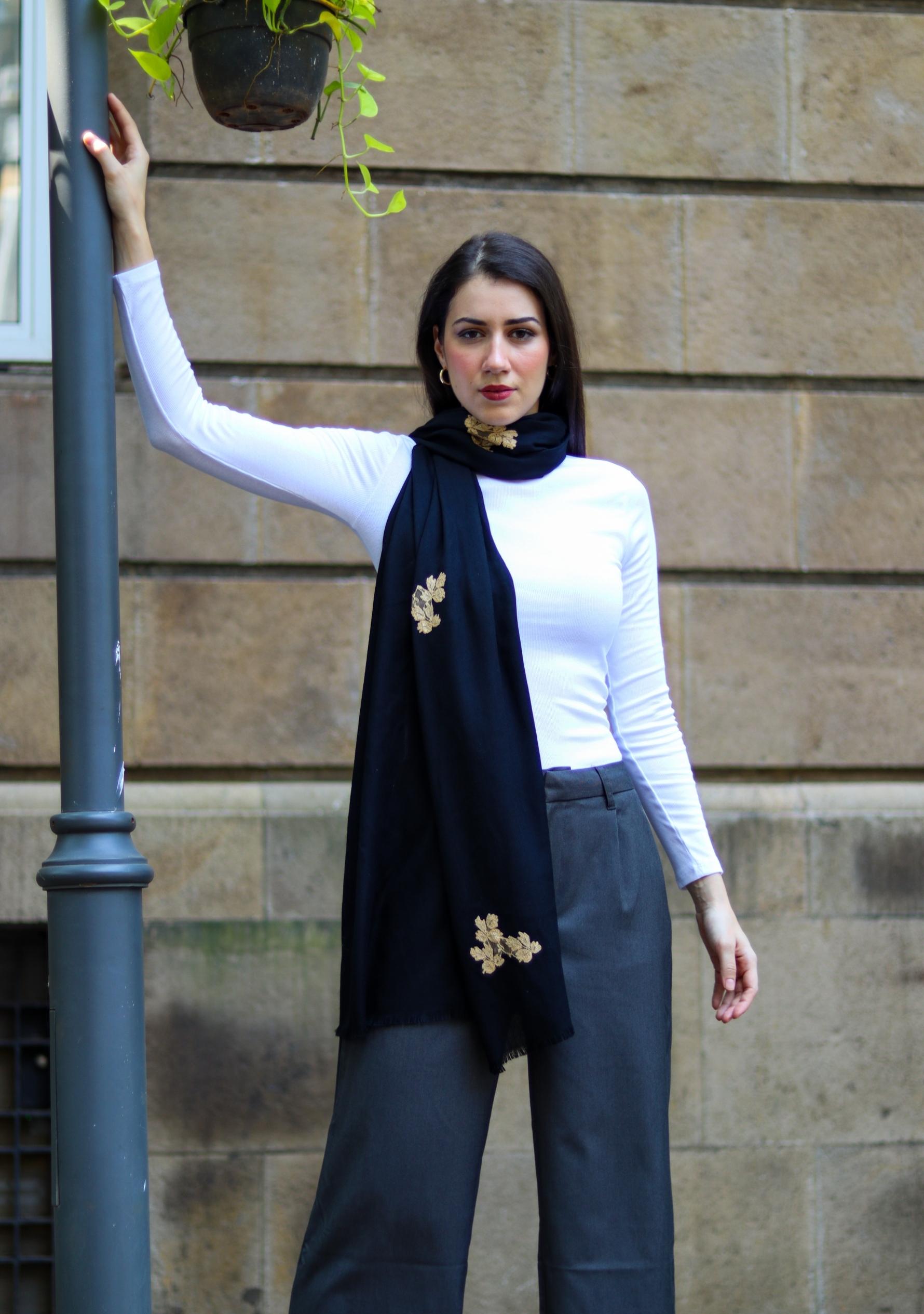 Woman wearing a white top and navy scarf with gold brooches against a stone wall.