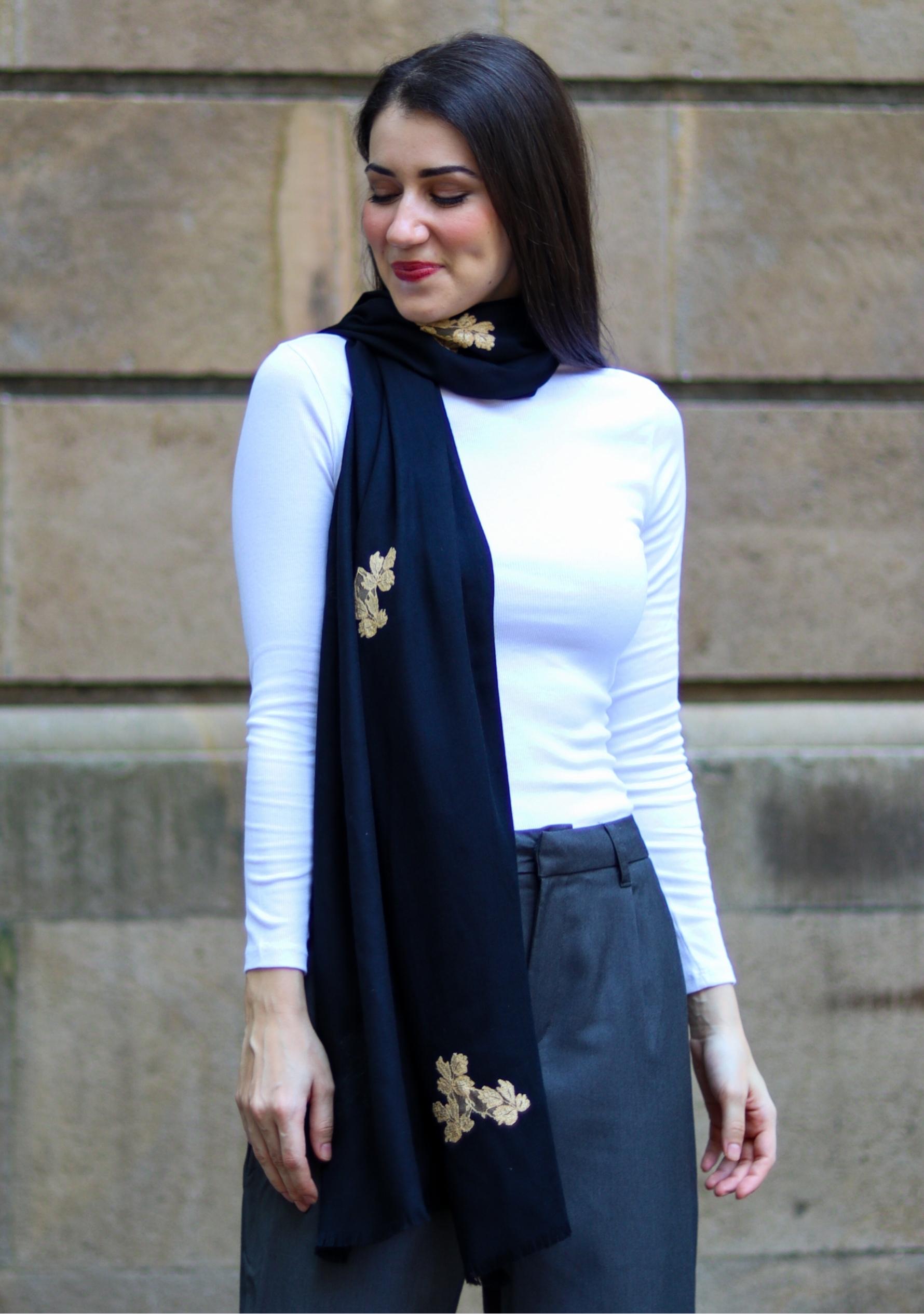 Woman wearing a black scarf with gold brooches against a stone wall.