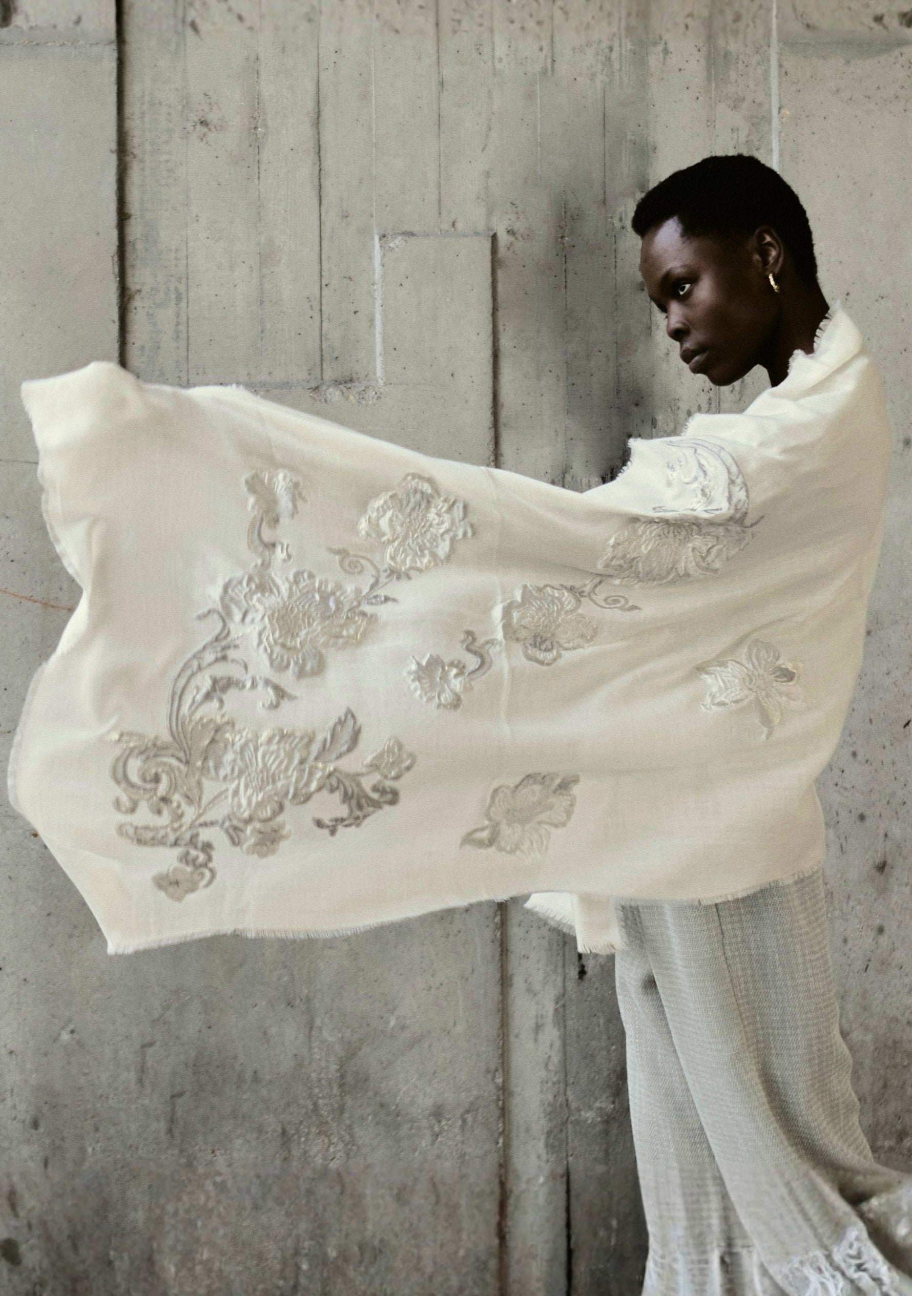 Person holding an embroidered white shawl against a concrete wall