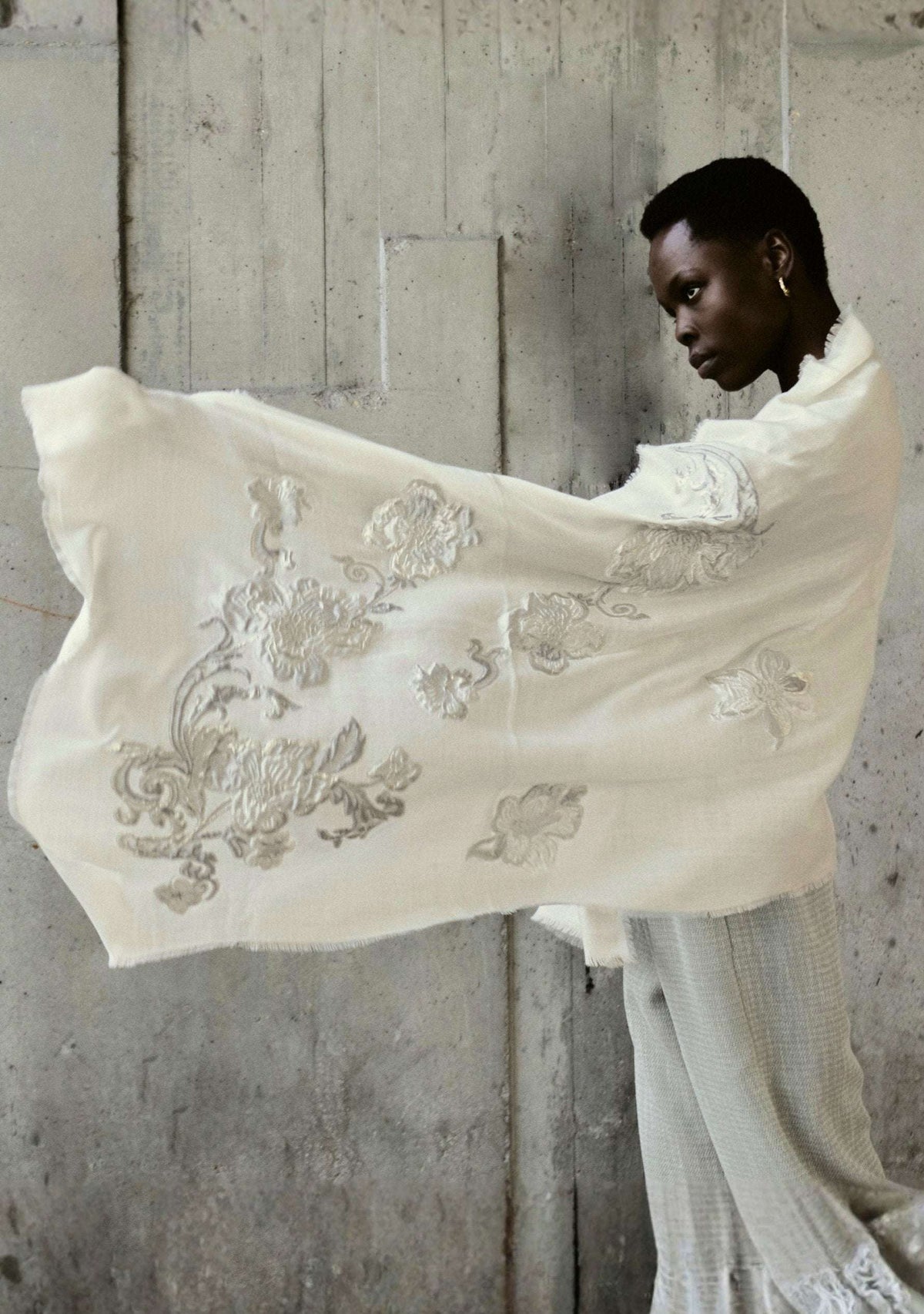 Person holding an embroidered white shawl against a concrete wall