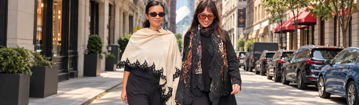 Two women walking side-by-side on a city street, showcasing black and beige fashion accessories including shawls and scarves.
