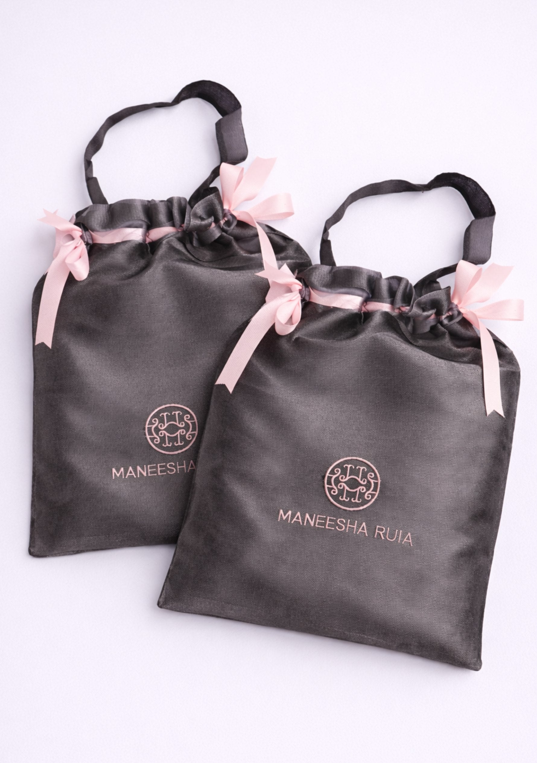 Two gray drawstring bags with pink ribbons and 'Maneesha Ruia' branding on a white background.