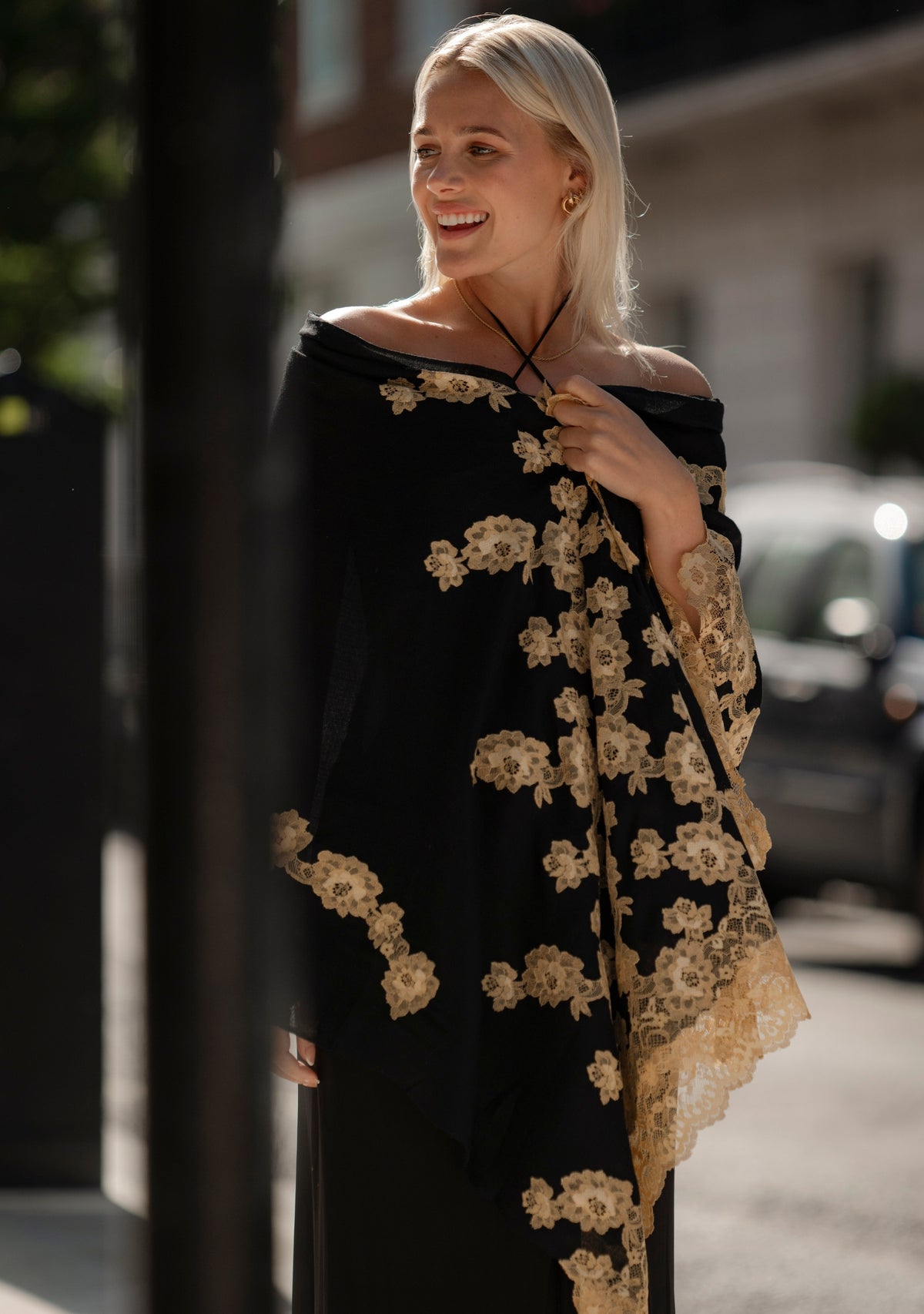 Luxury black cashmere scarf with handcrafted gold Chantilly lace in a floral vine pattern, elegantly draped on a smiling female model wearing a black off-shoulder dress. Soft, lightweight designer scarf with intricate lace detailing, perfect for formal occasions and timeless luxury styling.