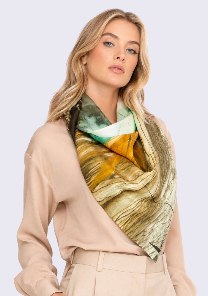 Grand Prismatic Print Silk Square Scarf by Maneesha Ruia elegantly draped around the neck, featuring a vibrant Yellowstone-inspired print in golden amber, emerald green, ivory, black, and earthy brown on luxurious 100% silk.