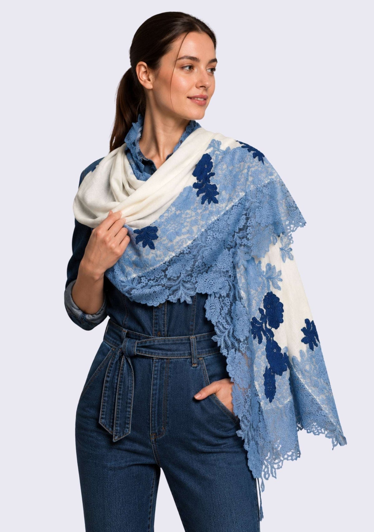 Female model wearing a luxury ivory wool and silk scarf with handcrafted steel blue and dark denim blue Chantilly floral lace, elegantly draped over a denim jumpsuit to showcase its lightweight fabric, flowing silhouette, and intricate floral lace craftsmanship.
