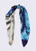 Mammoth Hot Springs Print Silk Square Scarf by Maneesha Ruia styled in a graceful knot, showcasing a scenic landscape print with vivid turquoise-blue skies, black accents, and white geothermal terrain on luxurious 100% silk.