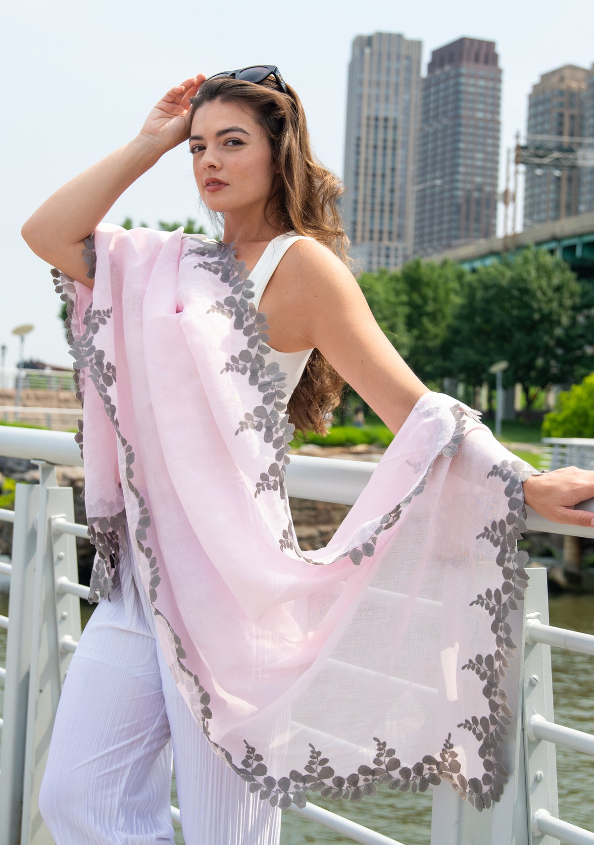 Woman holding a light pink scarf with gray floral patterns by a waterfront with buildings in the background