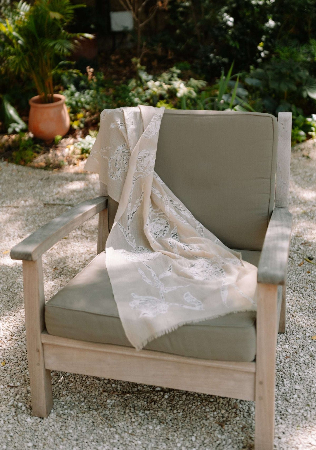 A light beige lace throw scarf with a floral pattern draped over the cushions of a wooden outdoor armchair on a gravel patio with green garden foliage in the background.