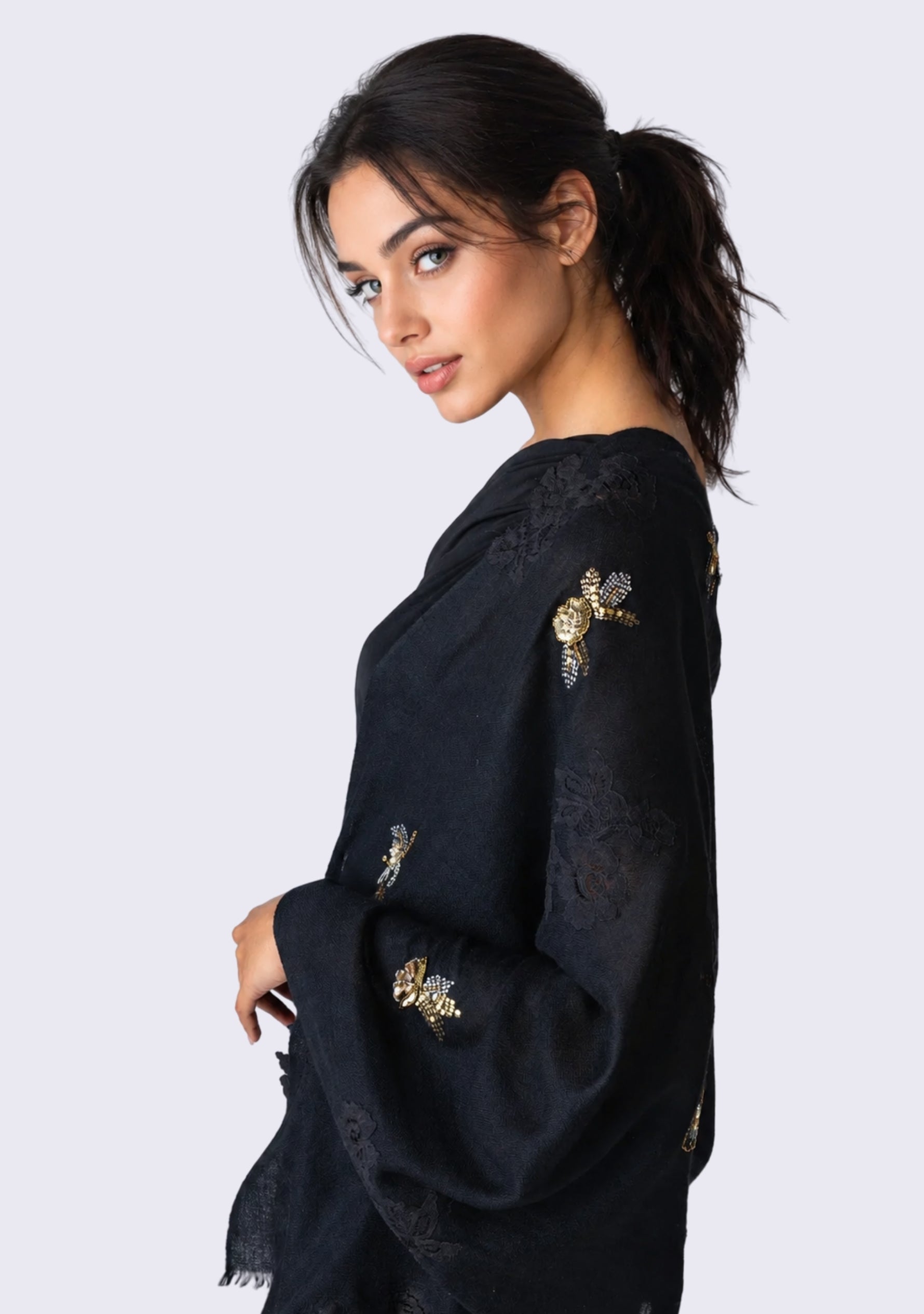 A woman wearing a black embroidered women's scarf with gold floral details, shown from a side-back angle against a light gray studio background.