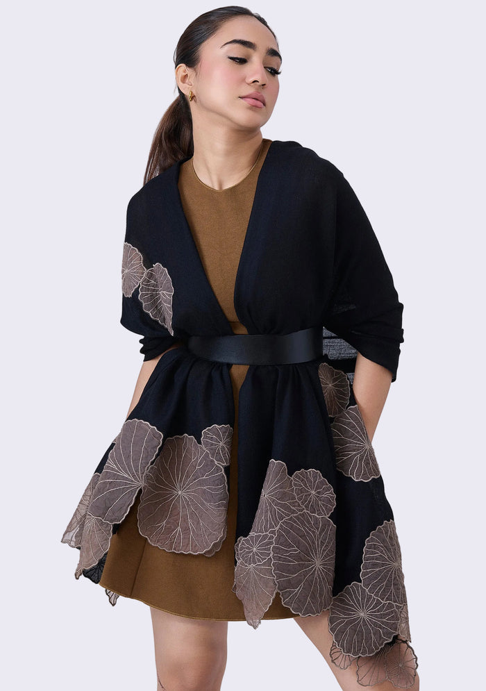 A woman wearing a black cashmere scarf with taupe lotus leaf appliqués, paired with a brown dress and a belt.