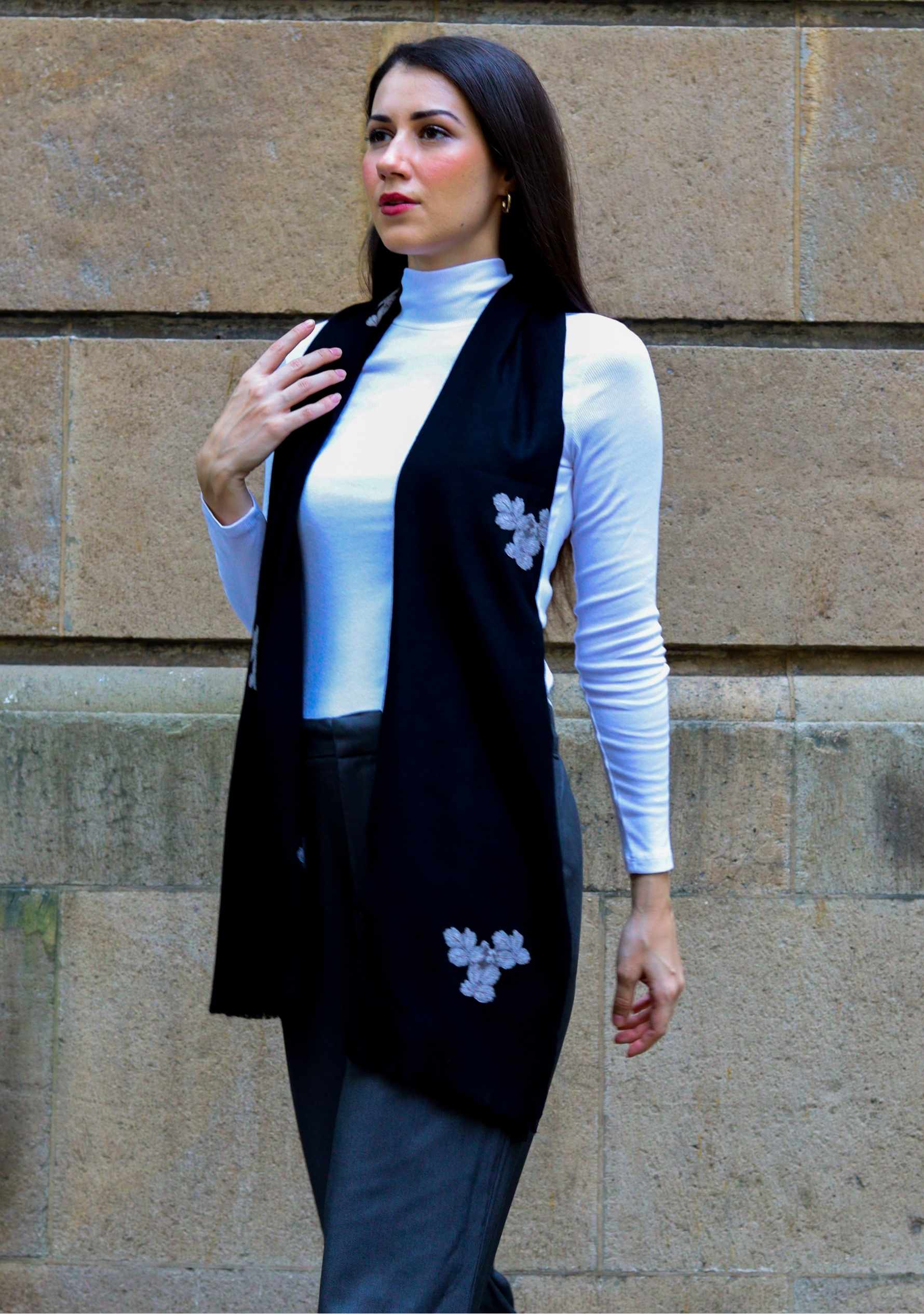 Woman wearing a black vest with floral embroidery against a stone wall.