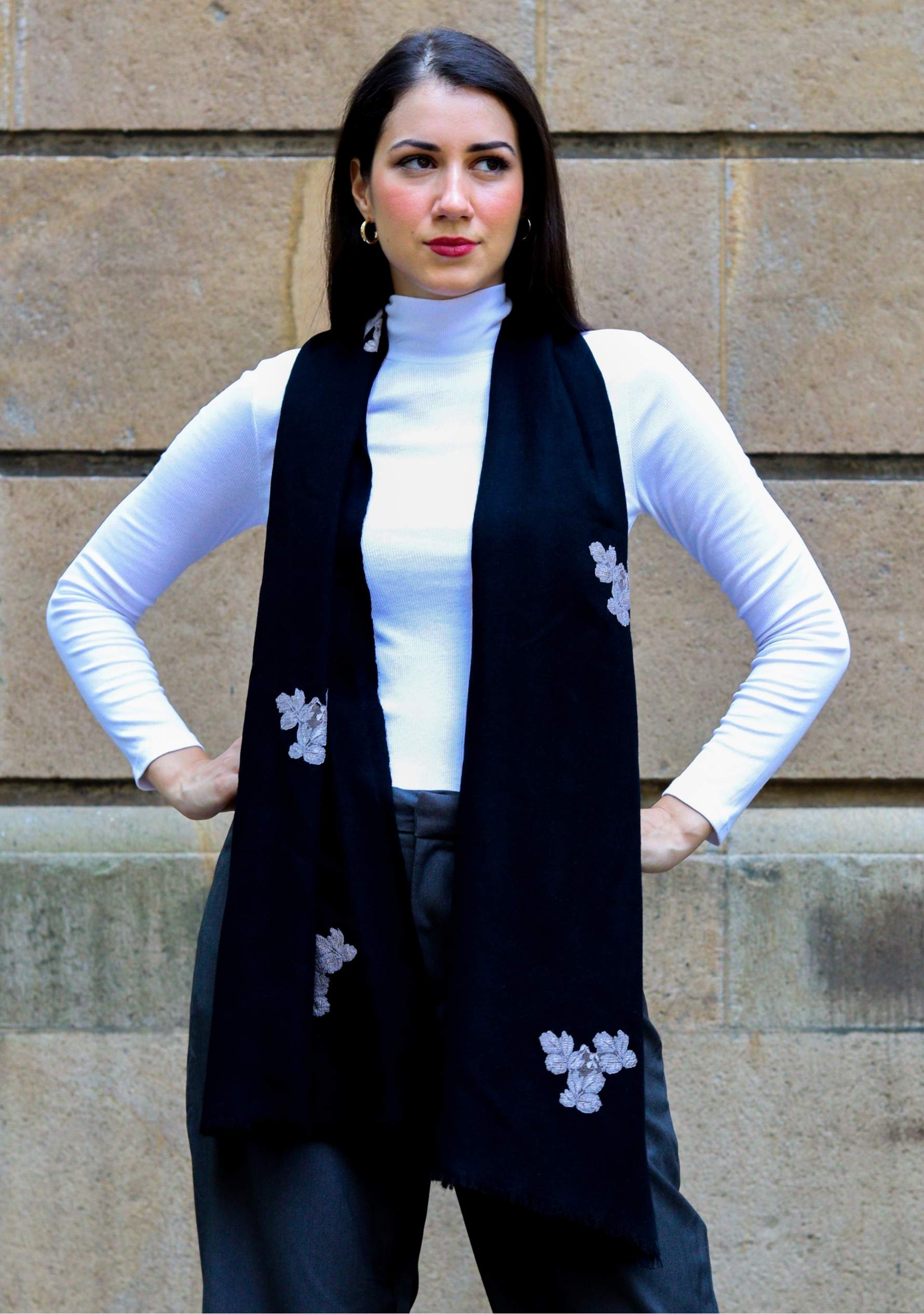 Woman wearing a black vest with white floral patterns against a stone wall.