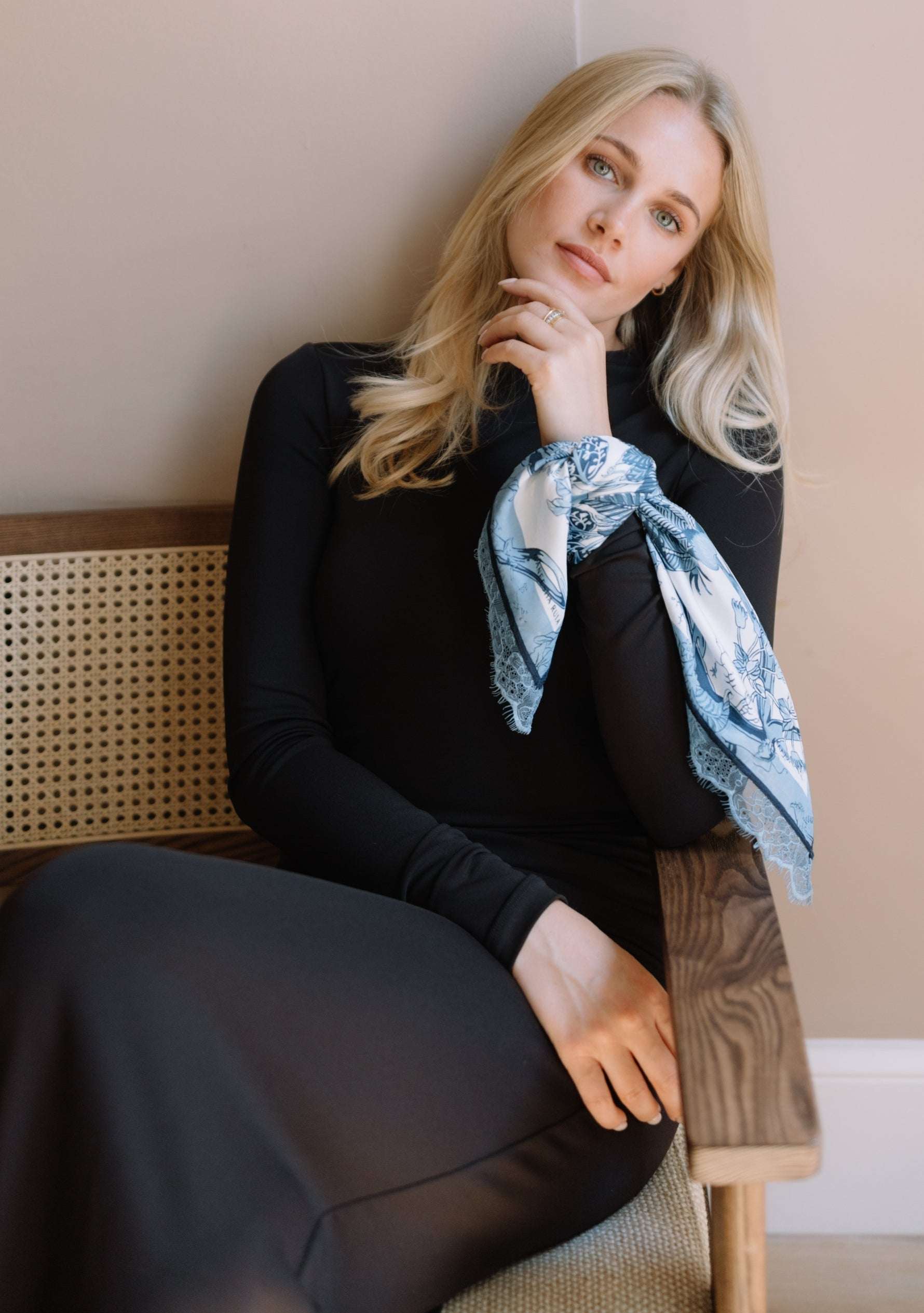 A blonde woman sits on a wooden chair with a cane back, wearing a black long-sleeve dress and a Maneesha Ruia's light blue patterned scarf tied around her upper arm.