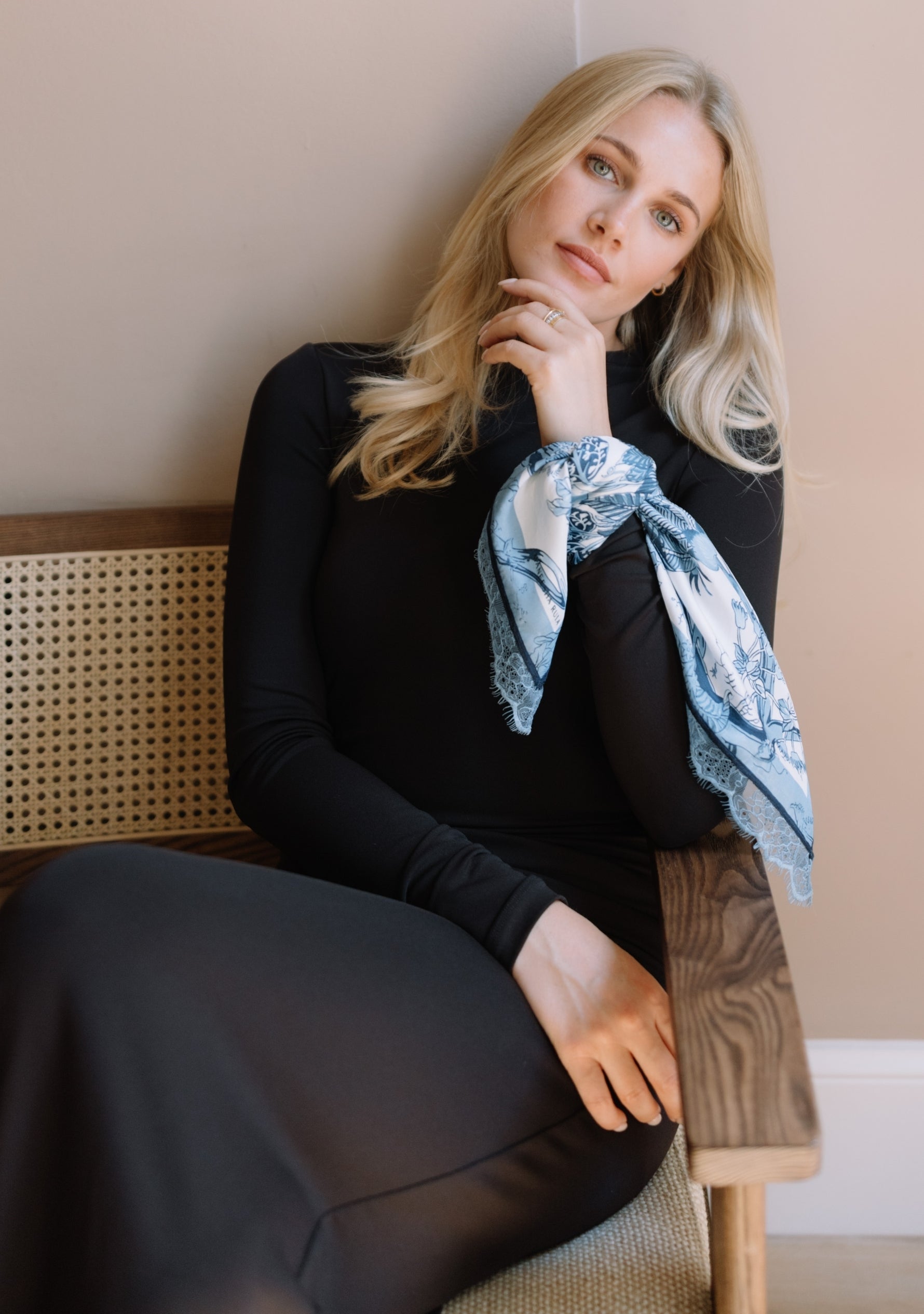 A blonde woman sits on a wooden chair with a cane back, wearing a black long-sleeve dress and a Maneesha Ruia's light blue patterned scarf tied around her upper arm.