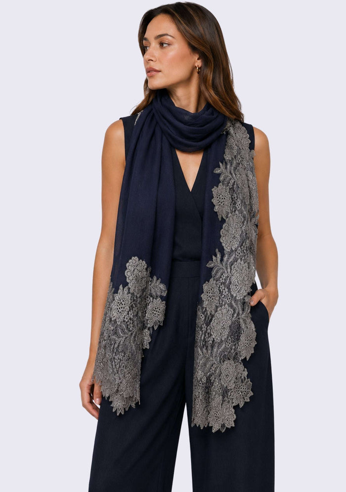 A woman modeling a dark denim blue sleeveless jumpsuit with a matching dark denim blue scarf featuring antique silver floral lace patterns, set against a plain light gray background.