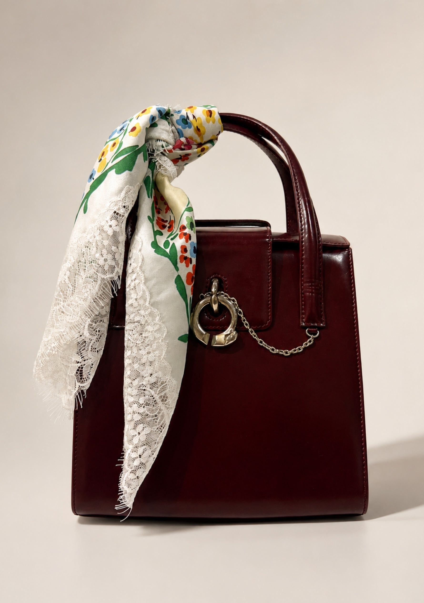 A structured burgundy leather handbag with a top handle, a silver-tone ring clasp, and a chain detail, styled with a white lace-trimmed scarf featuring a maneesharuia colorful floral print silk scarf.