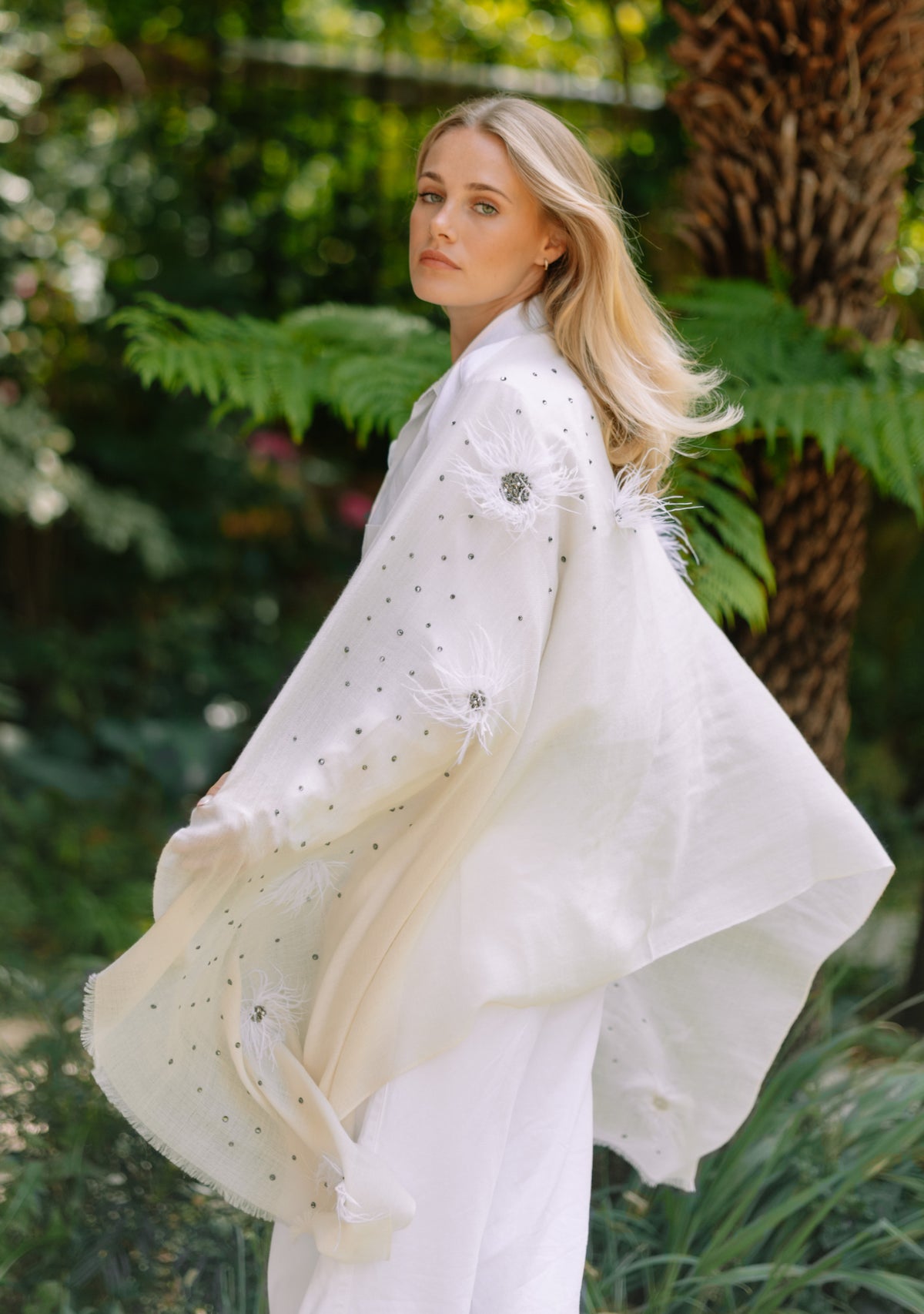 A woman wearing a flowing white embroidered Maneesha Ruia shawl or kimono-style wrap with sheer sleeves, photographed outdoors in front of lush green foliage and a tree trunk.