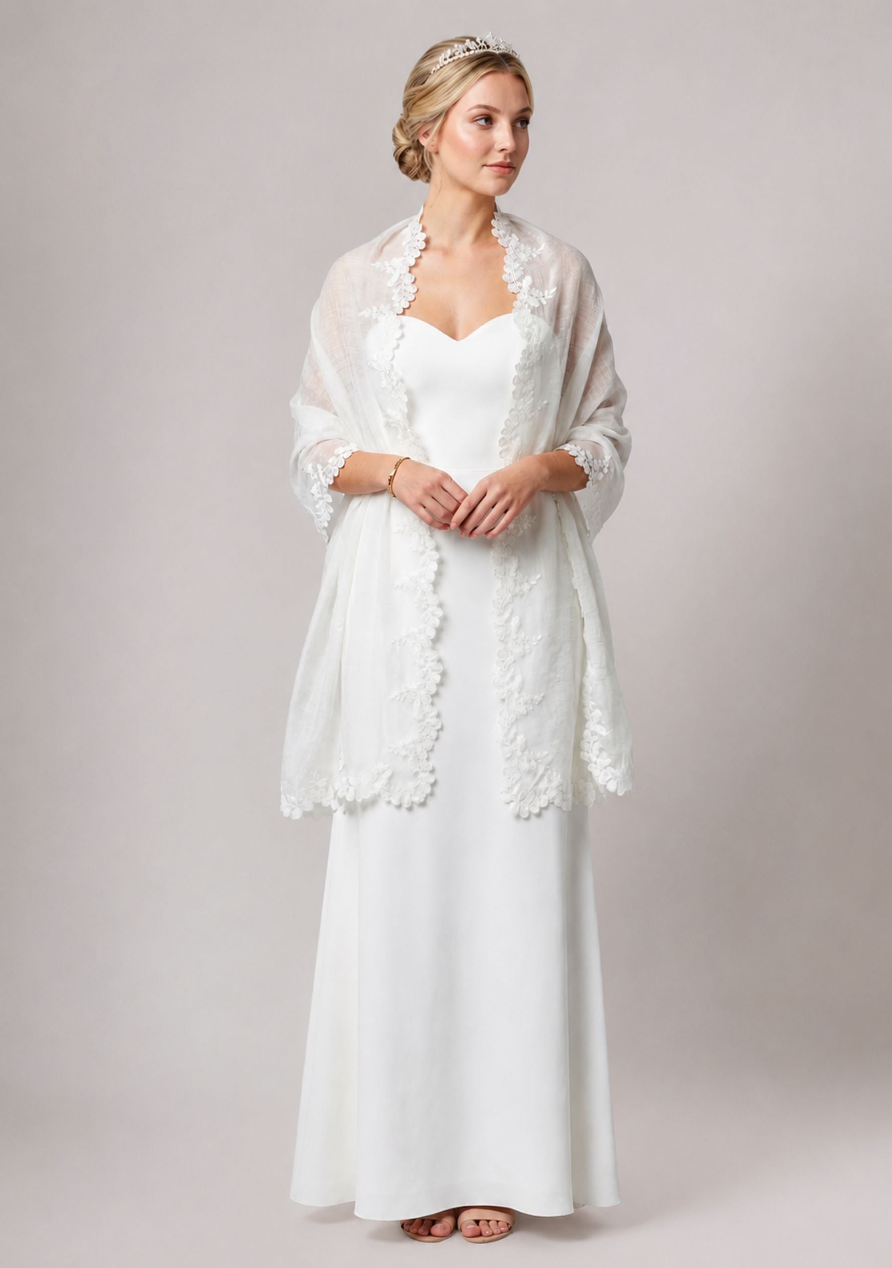 A woman modeling a white bridal gown with a sheer white lace shawl draped over her shoulders, photographed against a plain light gray studio background.