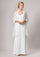 A woman modeling a white bridal gown with a sheer white lace shawl draped over her shoulders, photographed against a plain light gray studio background.