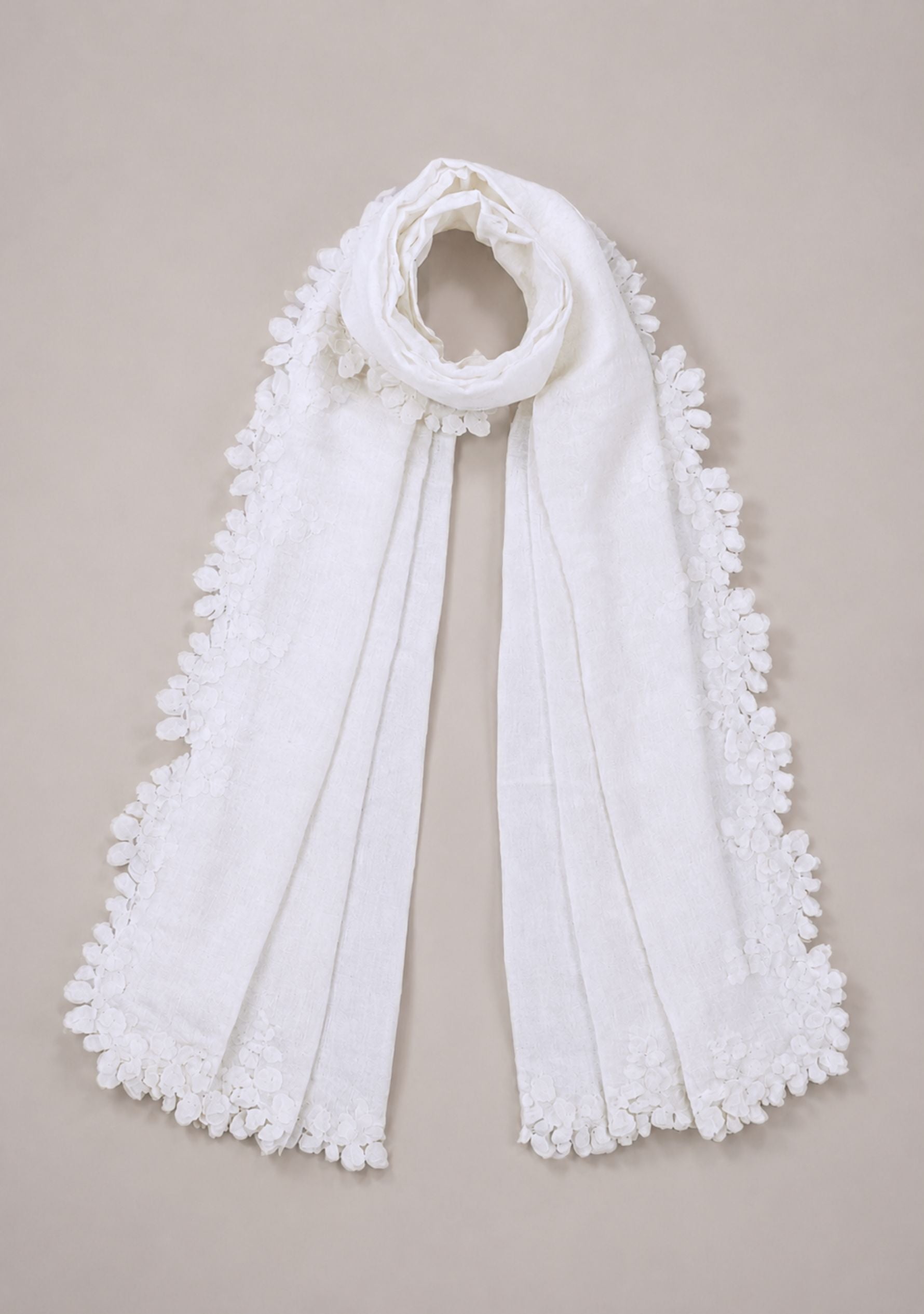 A white scarf with textured, raised floral or lace trim along the edges, draped in a loop at the neck on a solid beige background.