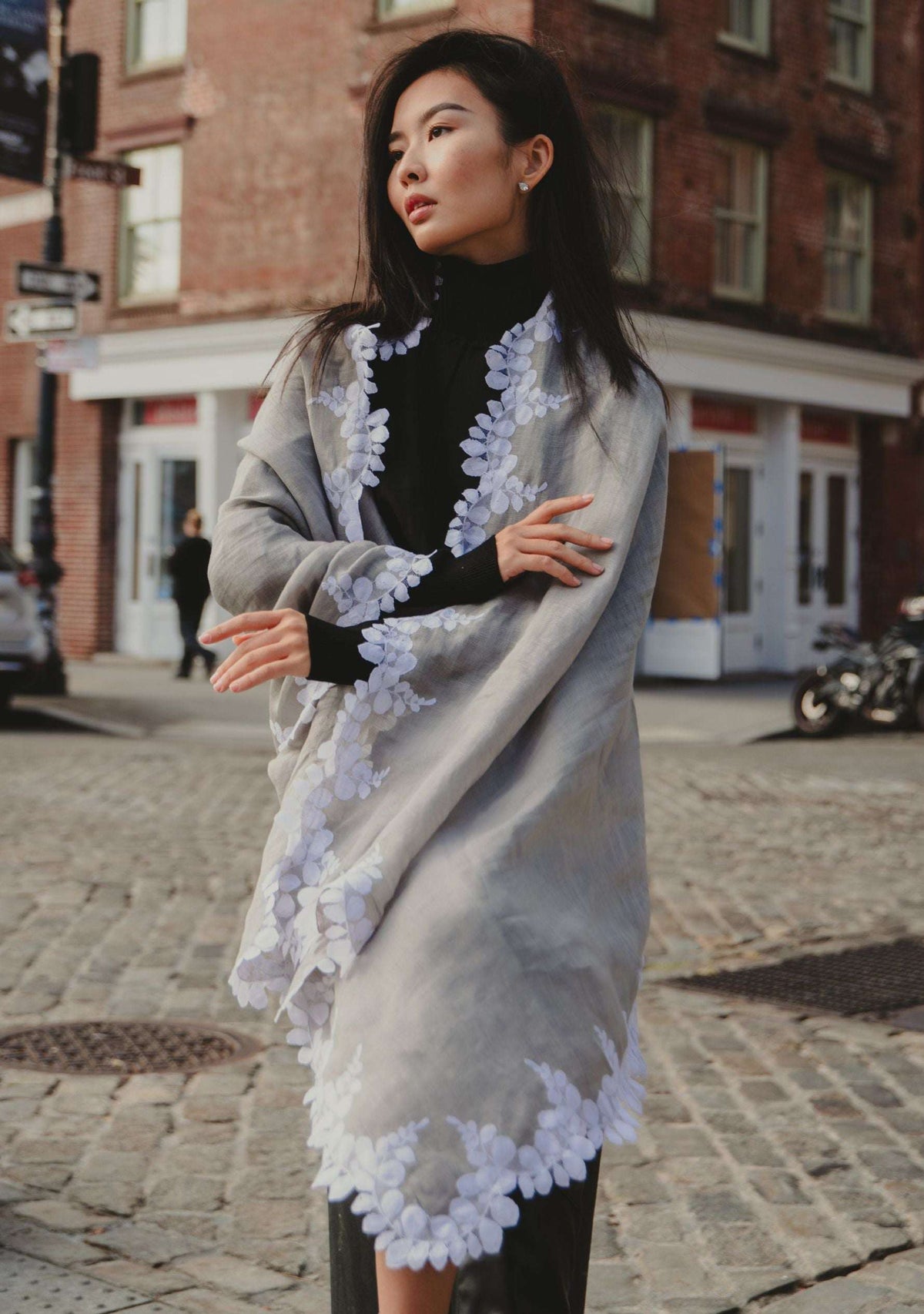 Woman wearing a light gray shawl with white floral patterns on a city street.