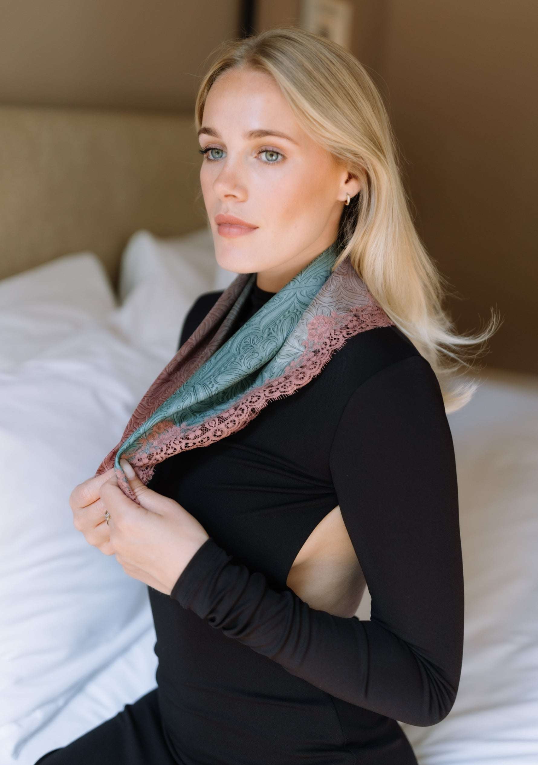 A blonde woman wearing a black long-sleeve top and a Maneesharuia sage Green - Pink ombre and dusty rose patterned scarf, sitting on a bed with white pillows.