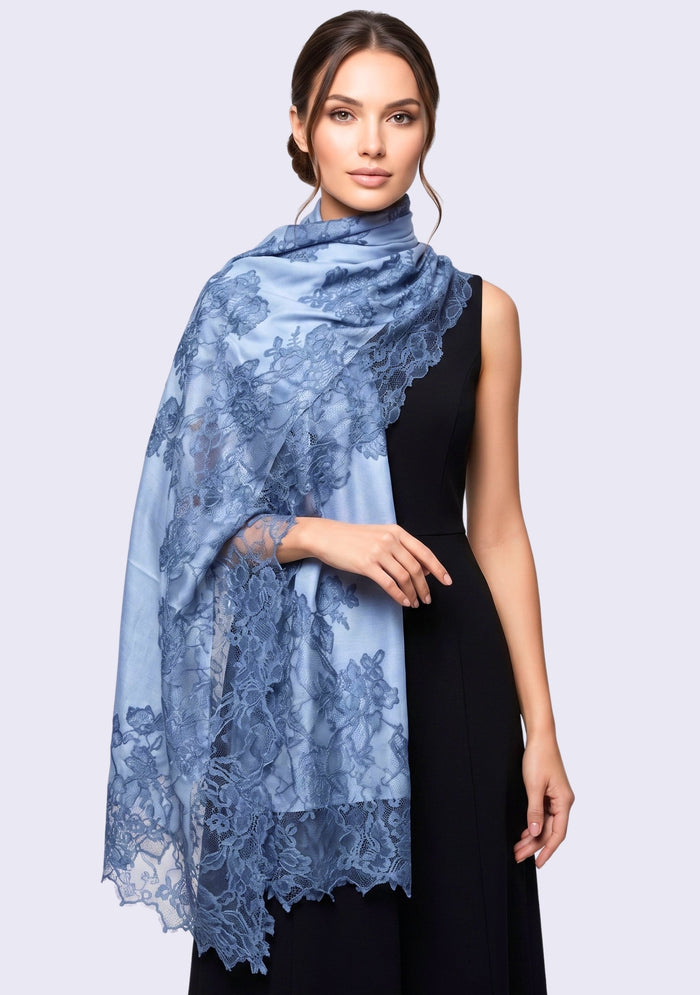 A woman modeling a light blue sheer lace shawl draped over her shoulders, paired with a black sleeveless dress against a light gray studio background.