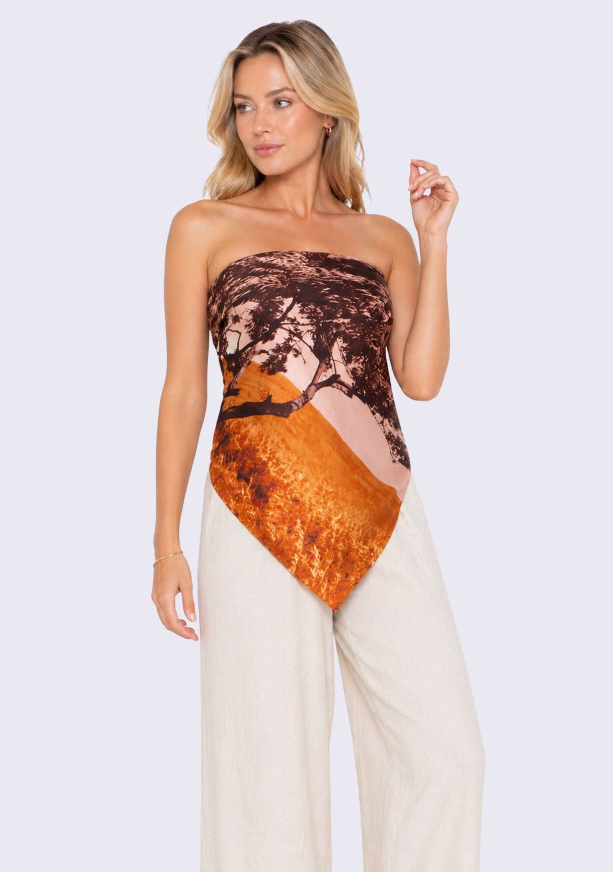 Santa Cruz Print Silk Square Scarf by Maneesha Ruia styled as a chic strapless silk top, featuring a scenic coastal print with warm rust orange, blush pink, deep brown, and black tones, paired with ivory wide-leg trousers for a luxury resort look.