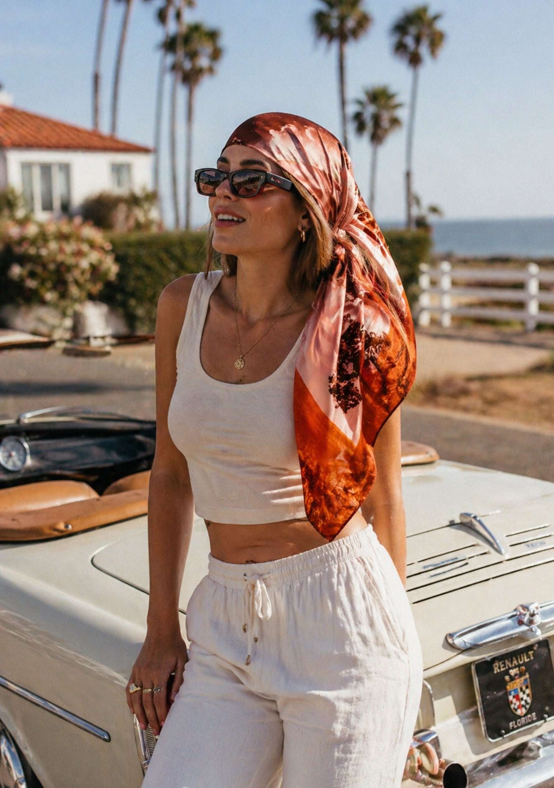 Santa Cruz Print Silk Square Scarf by Maneesha Ruia worn as a stylish headscarf, featuring warm coral, rust, blush pink, and deep brown coastal-inspired prints on luxurious 100% silk, paired with a white summer outfit.