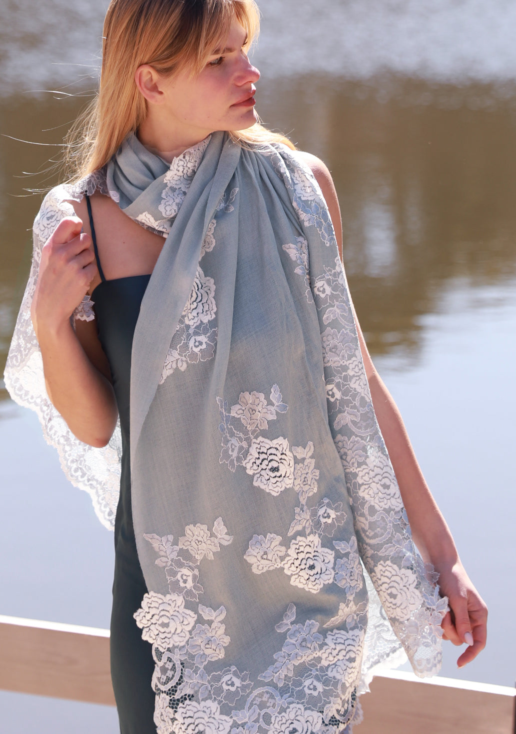 Luxury silver gray cashmere scarf with delicate light blue Chantilly floral lace, elegantly draped on a female model in a sleeveless dress. Soft, lightweight designer cashmere wrap with intricate handcrafted lace, perfect for weddings, evening wear, formal occasions, and timeless luxury styling.