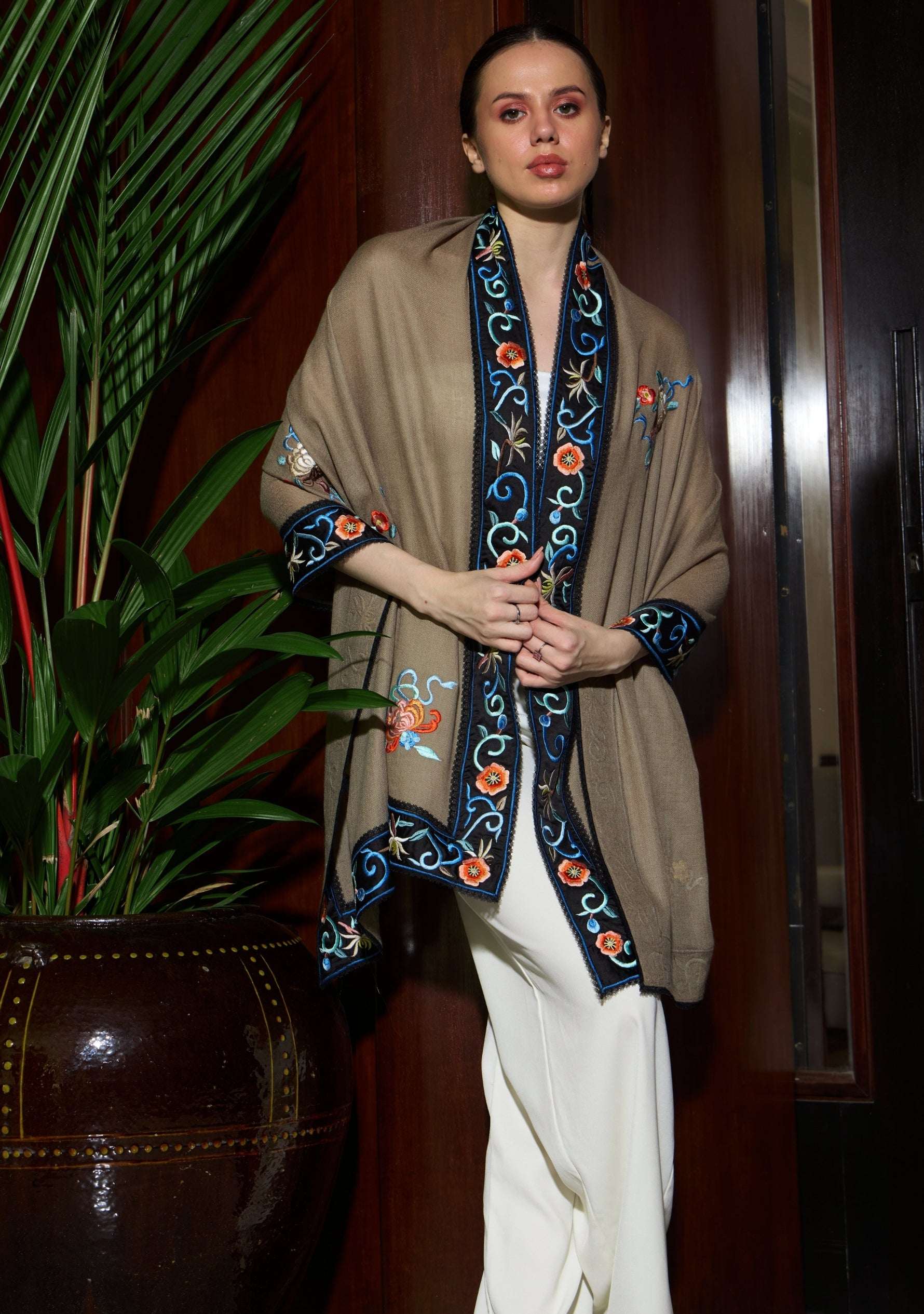 Woman wearing a beige shawl with intricate blue and orange patterns, standing indoors next to a plant.