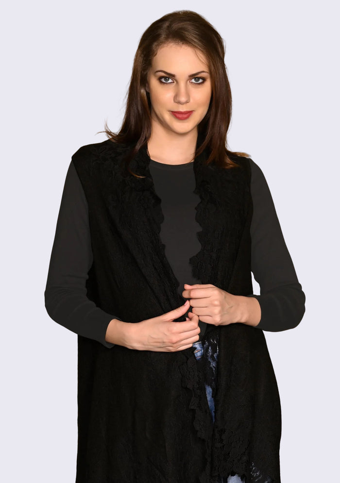 A woman wearing a black knitted sleeveless jacket with a floral lace border, over a black top and blue jeans.